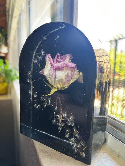 Decorative black plaque with a pressed rose and dandelion seeds on a windowsill.